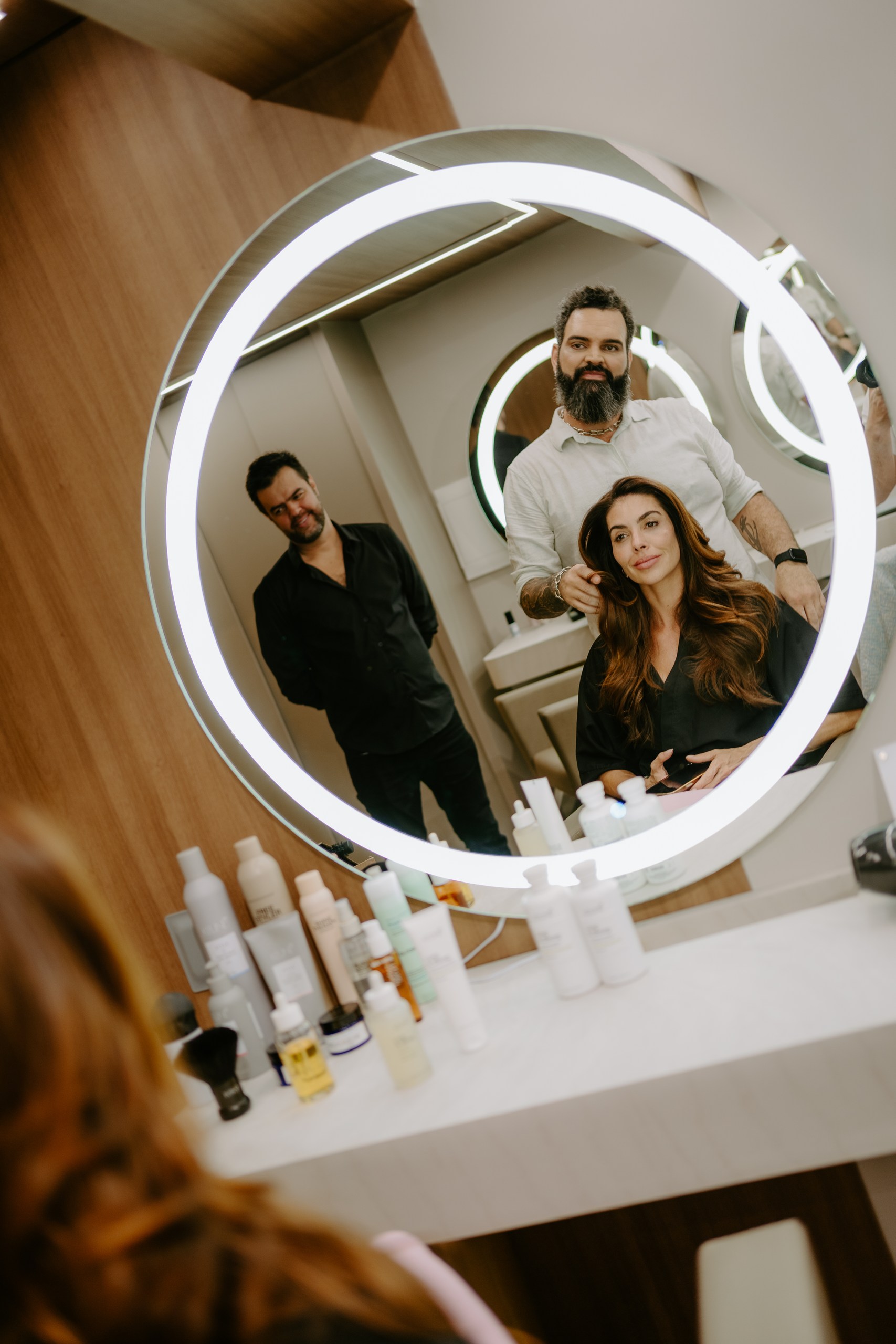 Felipe Messias finaliza o penteado de uma cliente, refletidos no espelho iluminado
